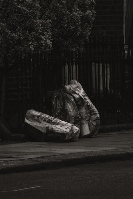 A narrow urban alleyway lined with brick and metal buildings, cluttered with large black and grey rubbish bags, some torn open, revealing discarded materials inside. In the foreground, a wheeled trash