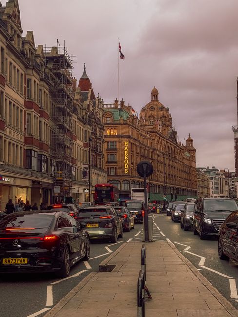 A busy street scene in Knightsbridge featuring a historic building with ornate architectural details and a prominent Harrods sign illuminated on the facade. The building has a large dome at the top an