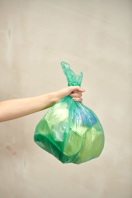 A pile of black rubbish bags and discarded items, including flattened cardboard and a wooden pallet, stacked against a weathered exterior wall beneath a red door covered with graffiti and stickers. Th